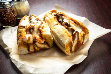 Chicken Parmesan hero sandwich cut in half, with breaded chicken cutlets, melted mozzarella cheese, and marinara sauce in a crusty hoagie roll, placed on parchment paper. Jars of grated Parmesan cheese and crushed red pepper flakes are in the background on a dark wooden table.