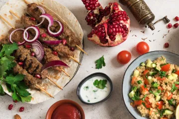 Middle Eastern meal with grilled meat kebabs on skewers garnished with red onion and pomegranate seeds, served on flatbread. Accompanied by a bowl of couscous salad with cucumber, tomato, and herbs. Additional garnishes include parsley, a halved pomegranate, cherry tomatoes, and small bowls of yogurt sauce and red sauce.