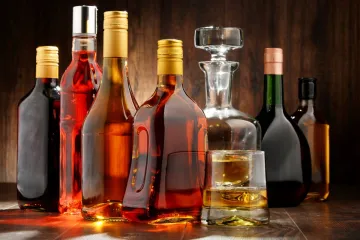 An assortment of spirit bottles in various shapes and colors on a wooden surface, with a glass of whiskey and a decanter.