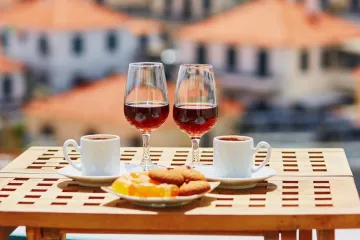 Two glasses of Madeira wine, two coffee cups, and a plate of sweets on an outdoor table overlooking blurred orange rooftops.