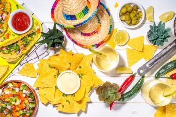 Overhead view of a festive Mexican-themed table with tortilla chips, tacos, salsa, lime drinks, peppers, sombreros, and colorful party decor.