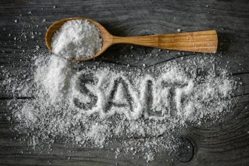 Wooden spoon filled with coarse salt on a dark table, with scattered salt spelling the word SALT.