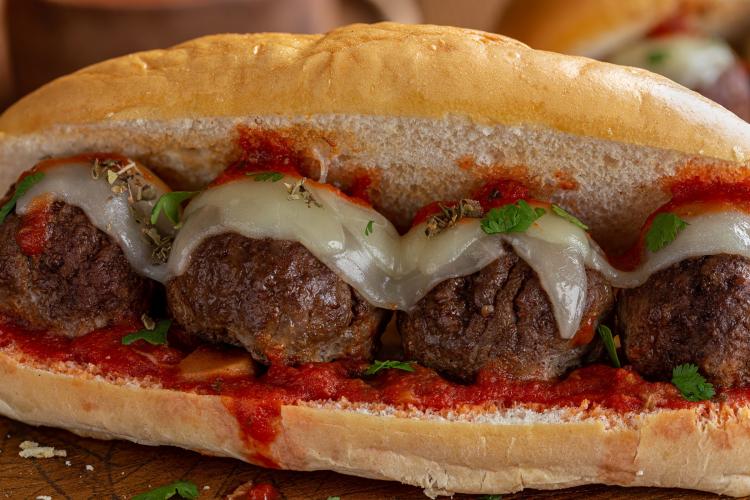 Close-up of a meatball hoagie sandwich with large meatballs topped with melted provolone cheese, garnished with parsley and dried herbs, in a soft hoagie roll with marinara sauce.
