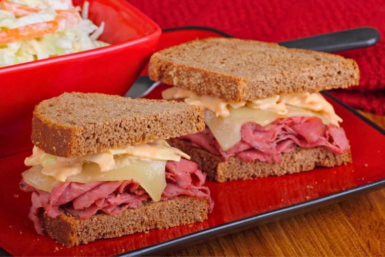 Two halves of a pastrami on rye sandwich with thinly sliced pastrami, melted Swiss cheese, and creamy coleslaw dressing on rye bread, displayed on a red plate. A bowl of coleslaw is visible in the background.