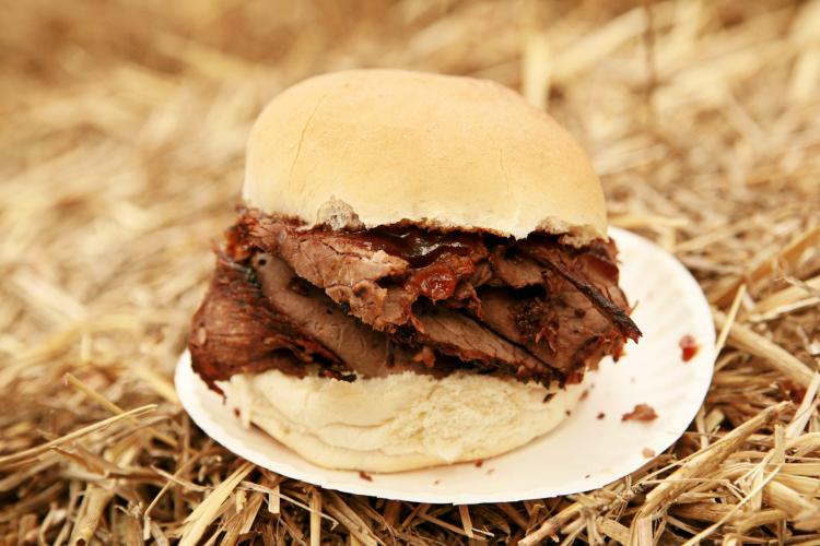 Pit beef sandwich with thick slices of smoked beef in a soft bun, placed on a white paper plate against a background of hay or straw.