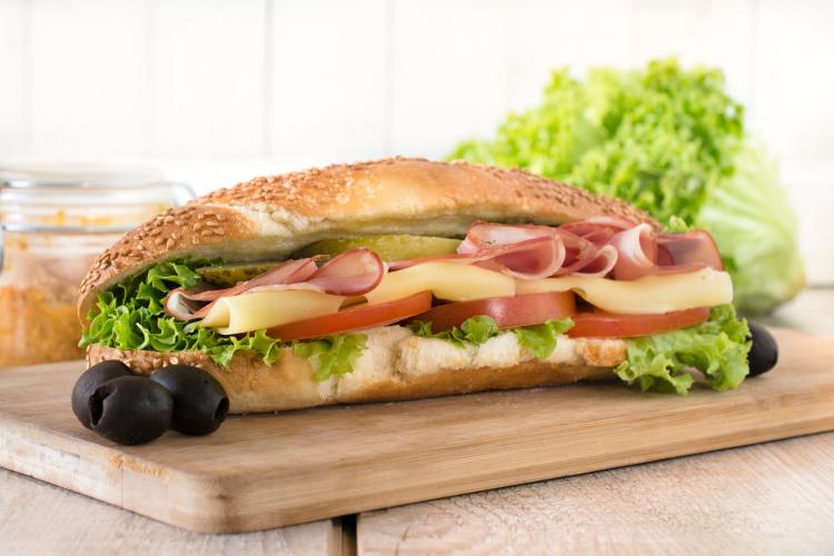 Submarine sandwich on a sesame seed hoagie roll filled with lettuce, tomato slices, yellow cheese, deli ham, and a pickle slice, placed on a wooden cutting board with two black olives beside it. Fresh lettuce and a jar of spread are in the blurred background.