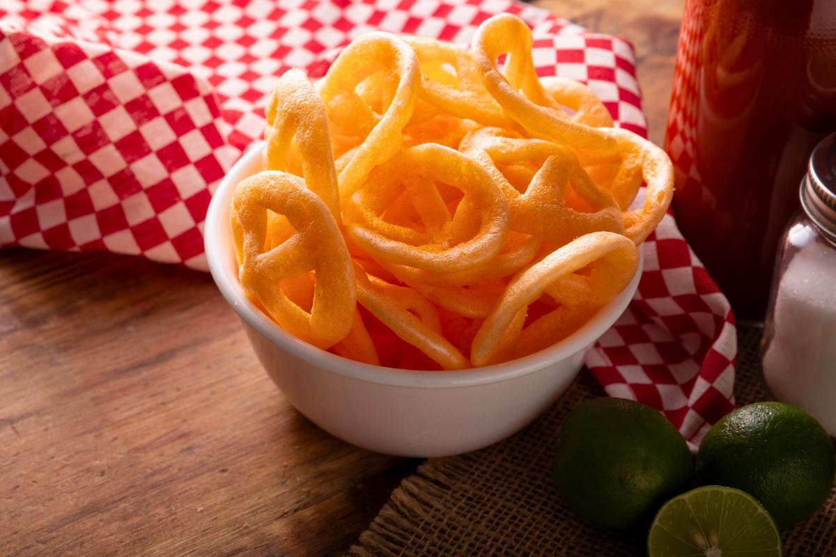 White bowl filled with crispy, golden, ring-shaped snacks on a wooden surface. Behind the bowl is a red and white checkered cloth. A glass container with a dark liquid, a salt shaker, and two whole limes along with a lime half are also visible.