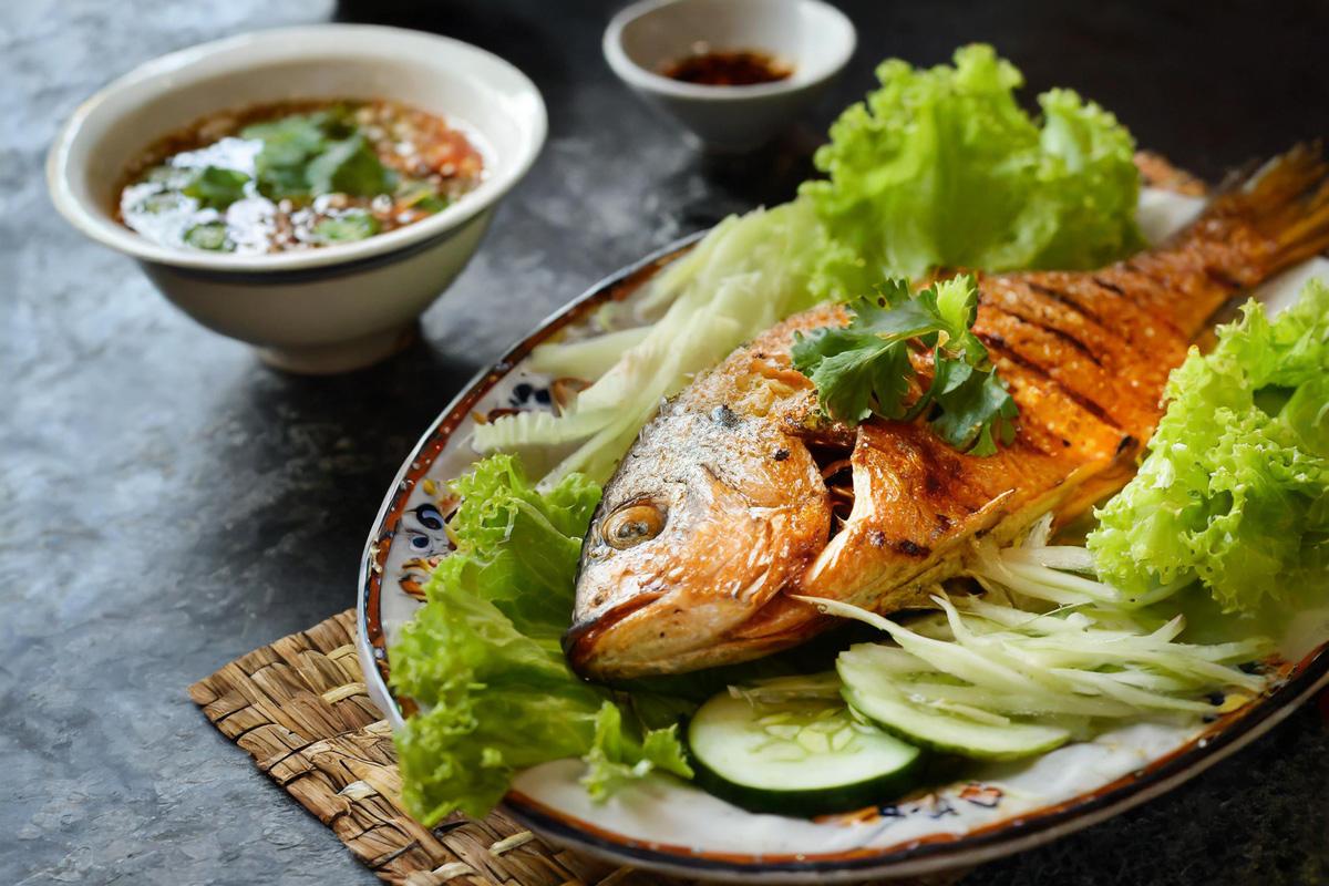 Vietnamese-style grilled fish served whole, garnished with cilantro and surrounded by lettuce and cucumber slices, with small bowls of dipping sauces in the background.