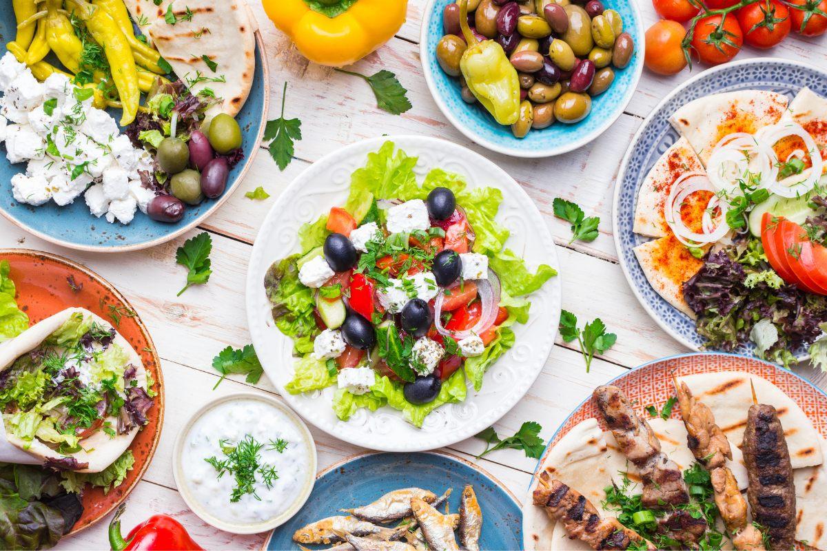 Top view of a Greek mezze platter featuring a Greek salad, pita bread, grilled meat skewers, olives, and mixed greens with feta cheese and peppers.
