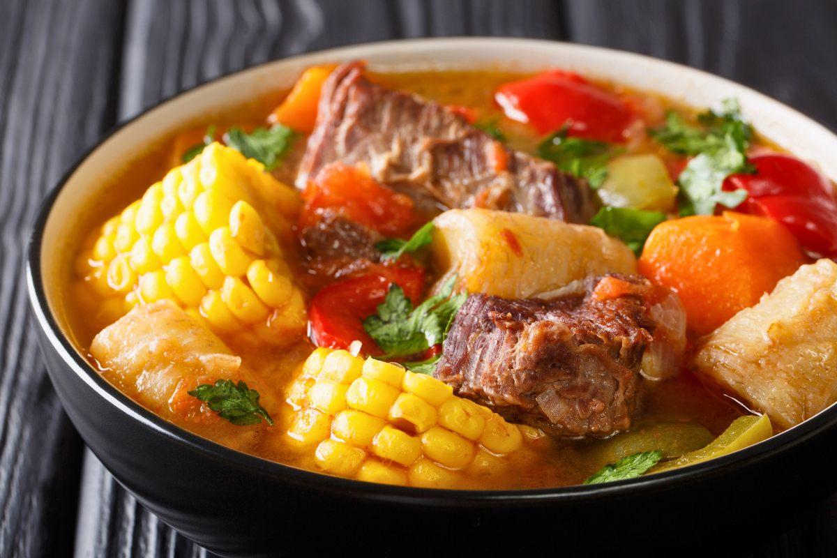A bowl of sancocho, a traditional Latin American stew, featuring beef, corn on the cob, root vegetables, and fresh herbs in a golden broth.