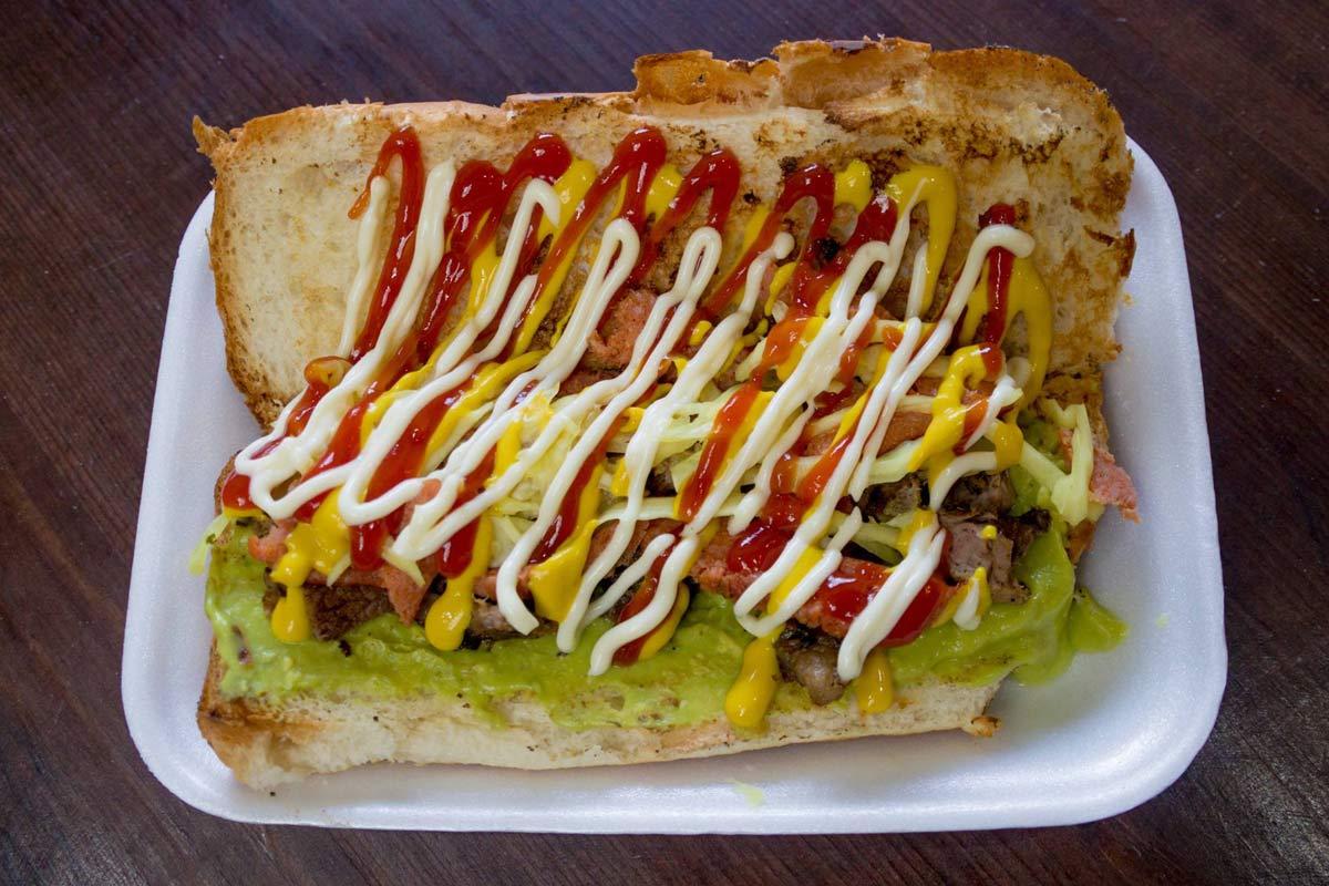 Guatemalan shuco hot dog topped with guacamole, cabbage, meat, ketchup, mustard, and mayonnaise on a foam plate.