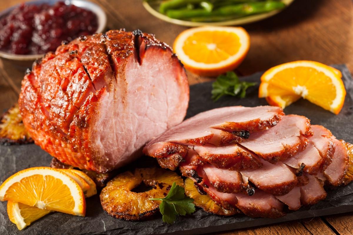 Sliced glazed baked ham served with orange wedges and grilled pineapple rings on a dark serving board.