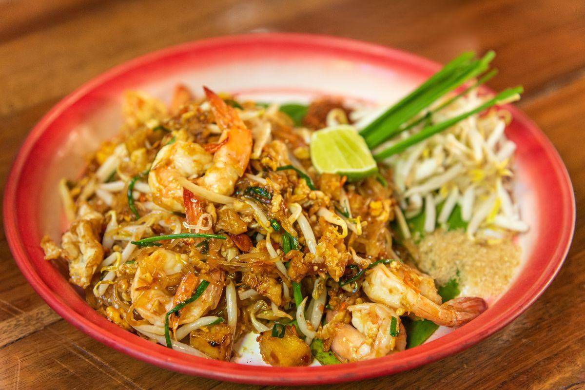 A plate of shrimp pad thai garnished with fresh bean sprouts, a lime wedge, and garlic chives, served on a red plate on a wooden table.