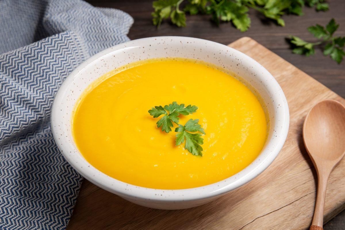 Bowl of potage Crécy, a creamy carrot soup, garnished with parsley on a wooden board beside a spoon and blue cloth.