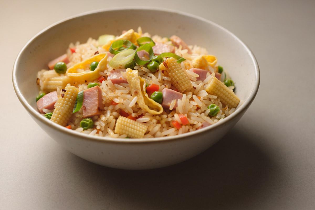A bowl of fried rice with diced ham, egg ribbons, baby corn, peas, carrots and scallions, on a warm beige background, synthetic image.