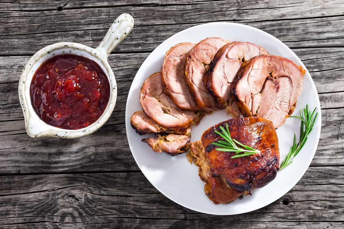 Sliced turkey roulade fanned out on a white plate with fresh rosemary, served alongside a small pitcher of chunky red chili-tomato jam on a rustic wooden table.