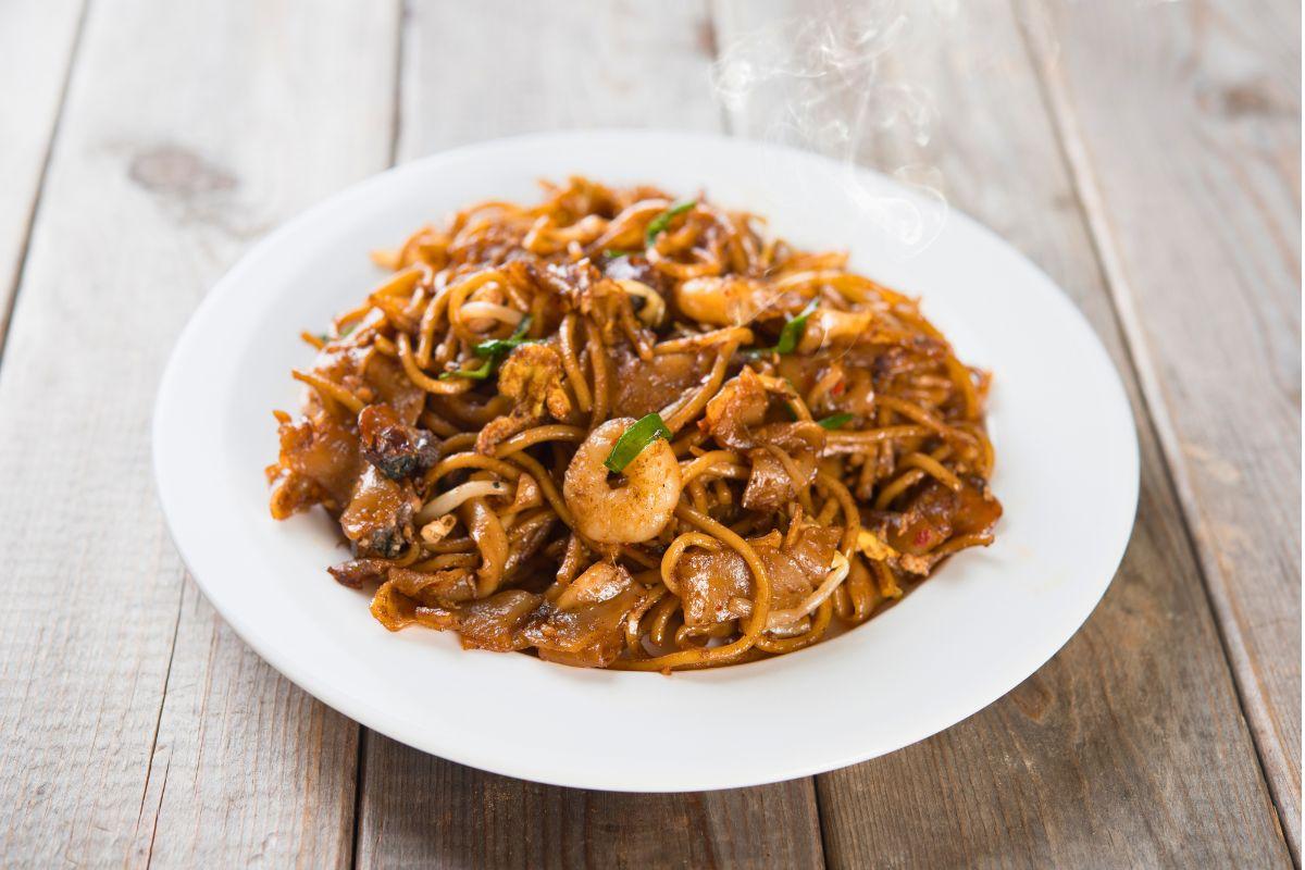 A plate of char kway teow — stir-fried flat rice noodles with shrimp, egg, bean sprouts and scallions, glistening with dark soy sauce and lightly charred, served on a white plate on a wooden table, with steam rising.