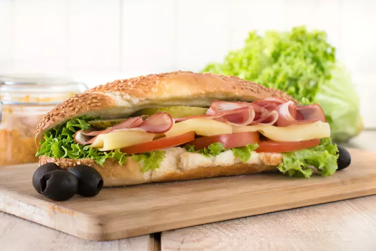Submarine sandwich on a sesame seed hoagie roll filled with lettuce, tomato slices, yellow cheese, deli ham, and a pickle slice, placed on a wooden cutting board with two black olives beside it. Fresh lettuce and a jar of spread are in the blurred background.