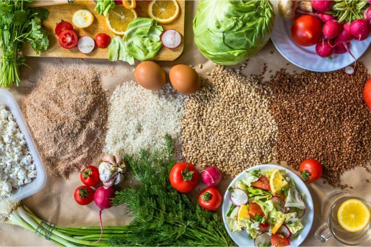 A colorful assortment of organic fresh vegetables, grains, eggs, cheese, and a salad arranged on a surface, including cabbage, tomatoes, radishes, and various herbs