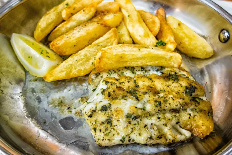 Grilled kingklip fish seasoned with herbs, served alongside thick-cut golden fries and a lemon wedge on a metal plate.