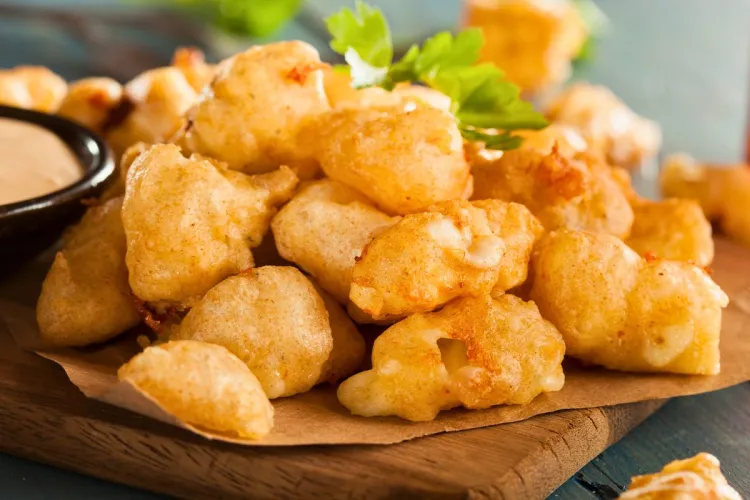 Golden, crispy battered cheese curds served on a wooden board with parchment paper, accompanied by a creamy dipping sauce and garnished with fresh parsley.
