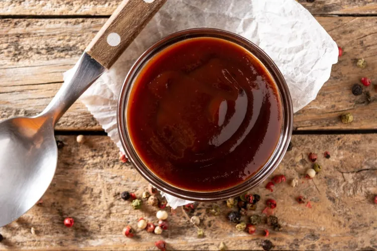 Close-up of a glass container filled with glossy barbecue sauce on a wooden surface, surrounded by scattered peppercorns and a spoon with a wooden handle.