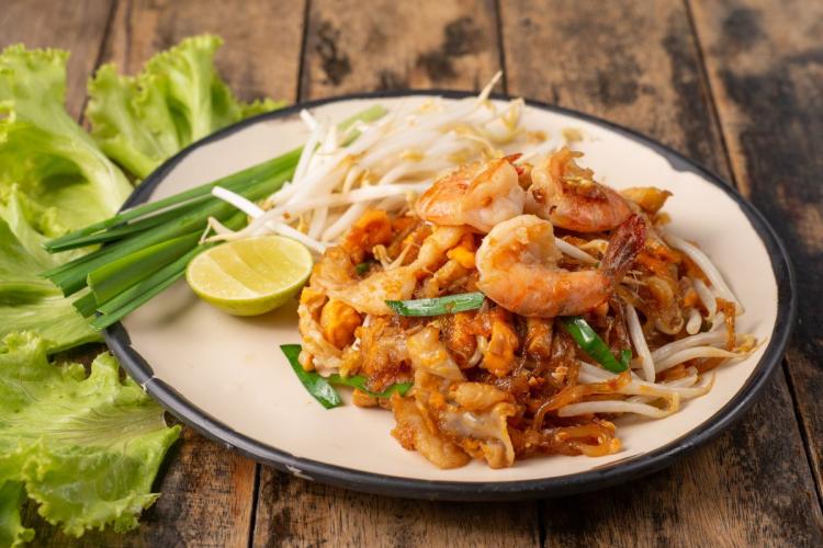 A plate of Pad Thai with shrimp, bean sprouts, green onions, and lime wedge on a rustic wooden table.