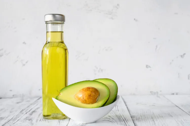 Bottle of avocado oil beside a halved avocado in a white bowl on a white table.