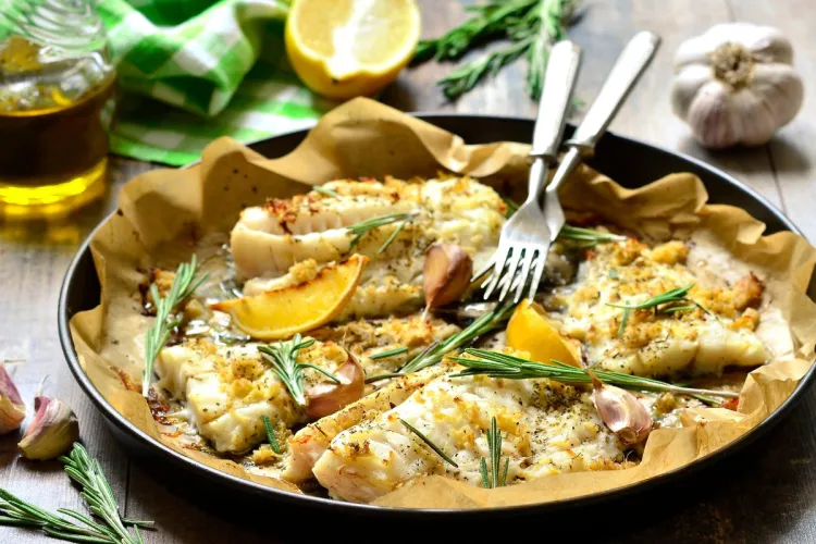 Baked cod fillets on parchment in a pan, topped with rosemary, garlic, and lemon wedges, with olive oil, fresh herbs, and a halved lemon in the background.