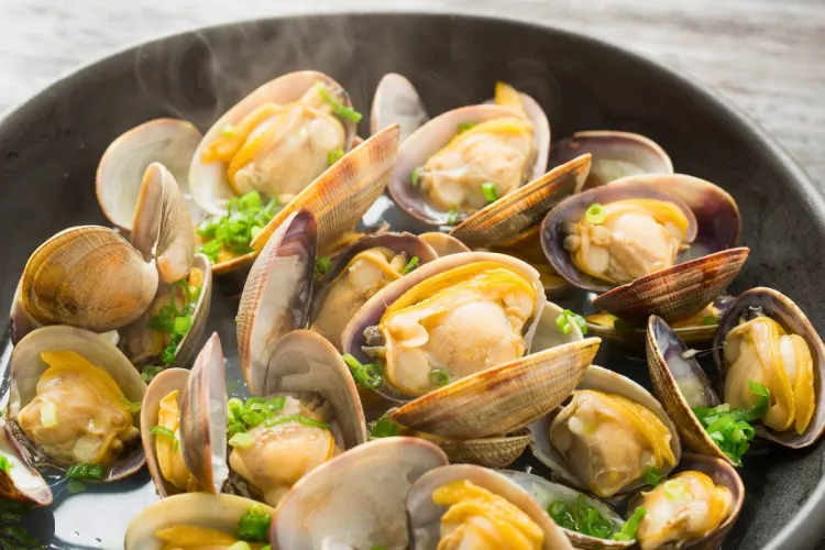 Close-up of a bowl of hot steamed clams in broth, garnished with chopped green onions and releasing steam.