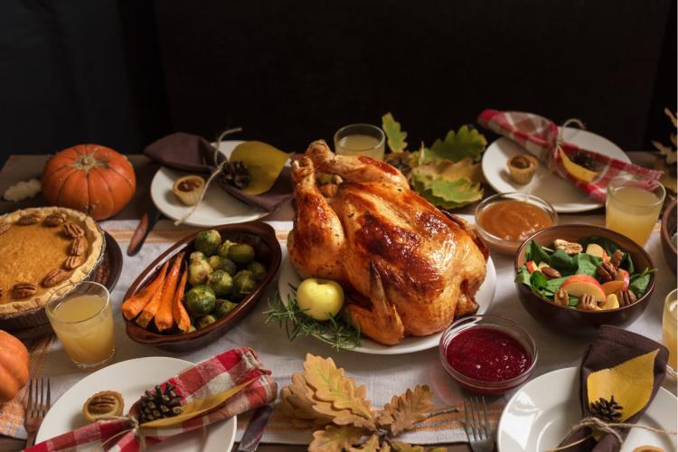 Holiday dinner table with a roasted turkey in the center, pumpkin pie, vegetables, salad, cranberry sauce, and fall decorations.