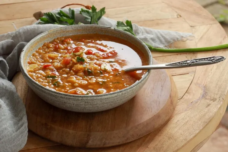 Speckled bowl of Greek lentil and vegetable soup with a spoon, set on a round wooden board with fresh parsley and a green onion nearby.
