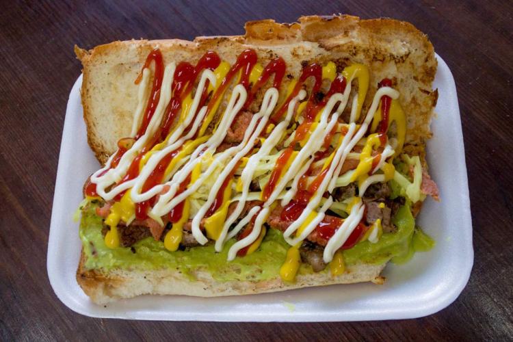 Guatemalan shuco hot dog topped with guacamole, cabbage, meat, ketchup, mustard, and mayonnaise on a foam plate.