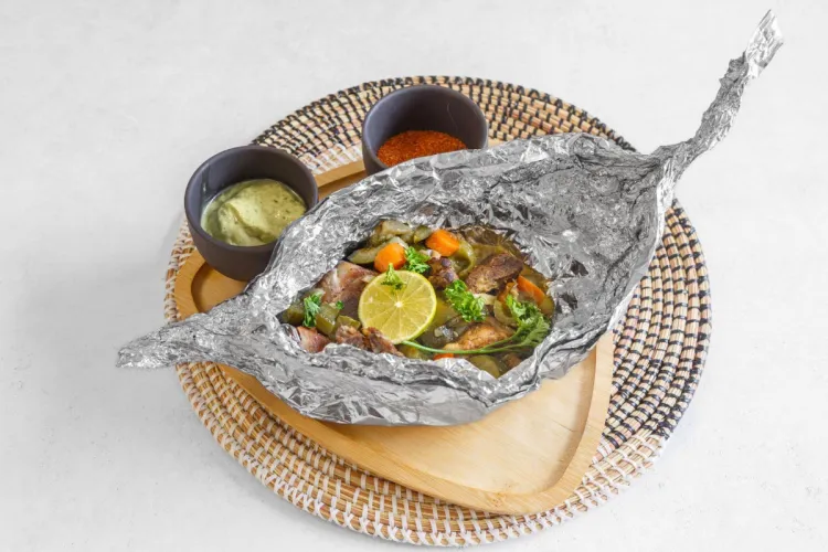Foil-wrapped fish with vegetables and lime served on a wooden tray with dipping sauces, Congolese style
