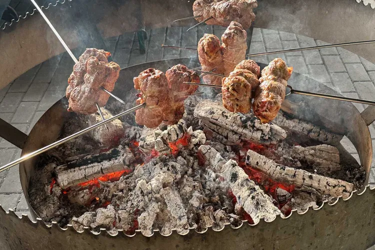 Several rolled meat roasts on metal spits cooking over glowing embers and burning logs in a large circular open-fire grill, with smoke rising from the coals.