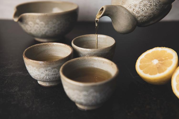 Amber tea being poured from a stoneware teapot into small ceramic cups with stamped white floral decoration, on a dark stone surface beside a halved lemon