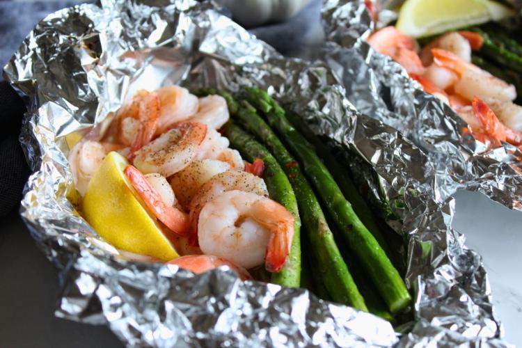 A foil packet filled with cooked shrimp, asparagus spears and lemon wedges, seasoned with black pepper.