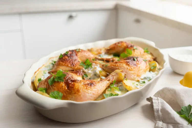 Baked chicken legs with sliced potatoes in a creamy lemon-yogurt sauce, garnished with fresh parsley, in a white oval baking dish.