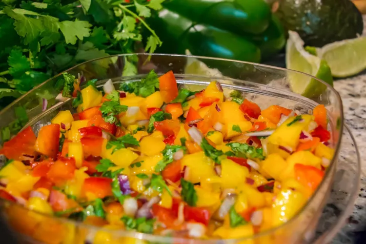 A bowl of tropical mango salsa with diced mango, red bell pepper, red onion and fresh cilantro, garnished with lime wedges, cilantro and a green bell pepper in the background.