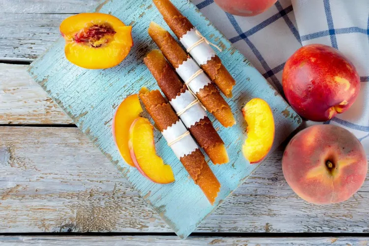Rolled strips of peach fruit leather tied with paper and string on a pale-blue board, with fresh whole peaches, a halved peach and slices