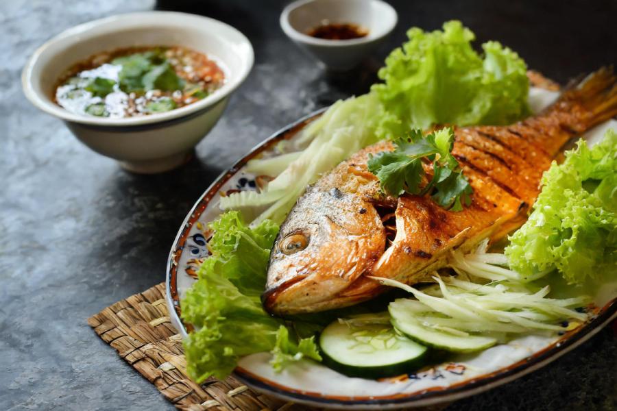 Vietnamese-style grilled fish served whole, garnished with cilantro and surrounded by lettuce and cucumber slices, with small bowls of dipping sauces in the background.
