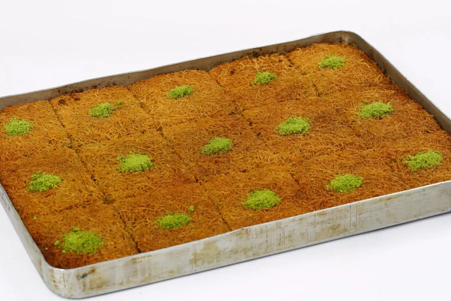 A rectangular baking tray filled with baked kataifi cut into squares, each topped with chopped pistachios.