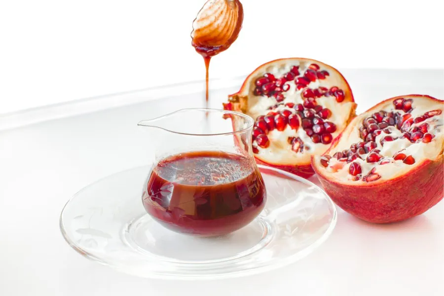 Glass jug filled with deep red pomegranate sauce on a decorative glass plate, accompanied by fresh pomegranate halves and a honey dipper dripping the sauce.