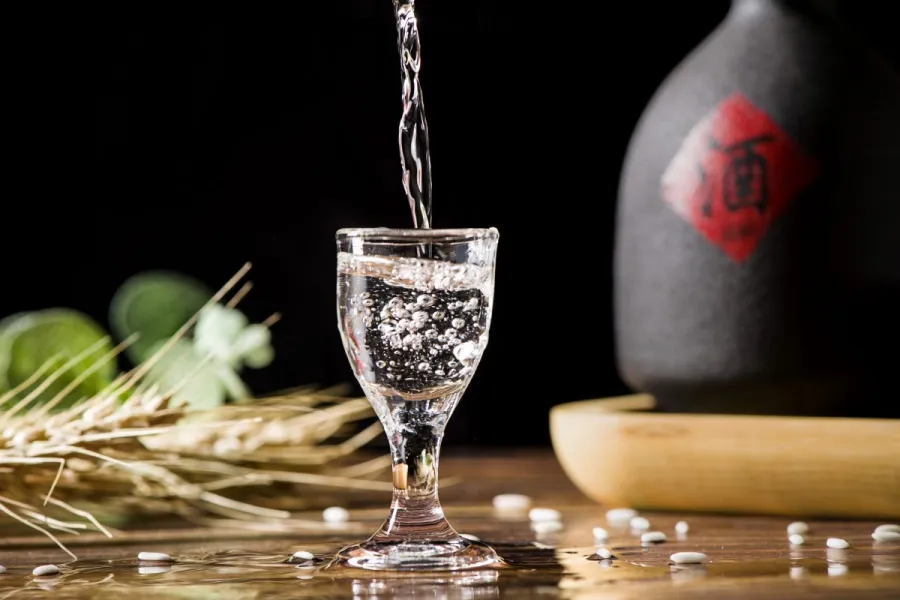 A clear bajiu being poured into a small glass with bubbles forming, with a ceramic bottle and scattered rice grains in the background.