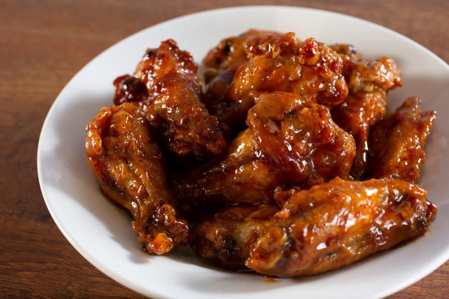 White bowl filled with glazed chicken wings coated in a thick, shiny, reddish-brown sauce, placed on a wooden surface.