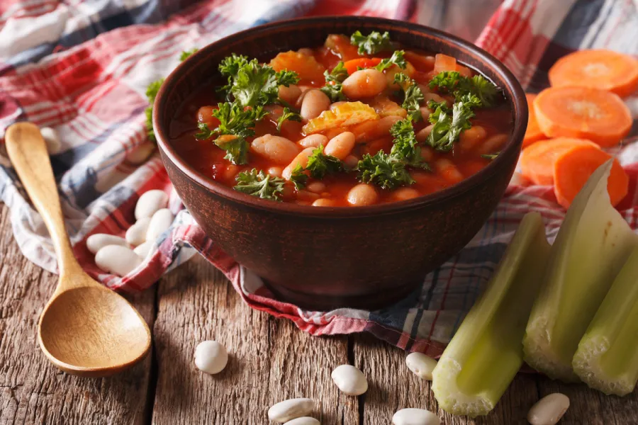 Bowl of traditional Greek Fasolada soup with white beans, carrots, celery, and parsley in a rich tomato broth, served on a rustic wooden table.