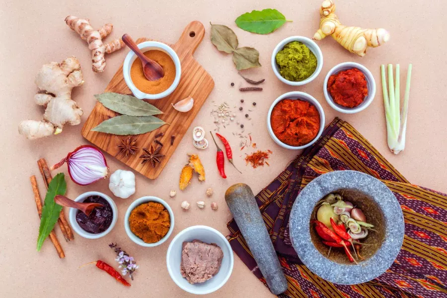 Flat lay of Thai cooking ingredients including fresh herbs, curry pastes, spices, and a stone mortar and pestle on a neutral background.