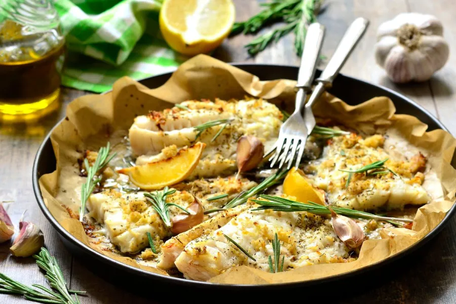 Baked cod fillets on parchment in a pan, topped with rosemary, garlic, and lemon wedges, with olive oil, fresh herbs, and a halved lemon in the background.