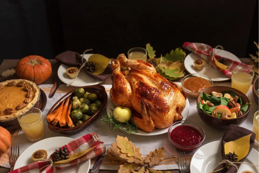 Holiday dinner table with a roasted turkey in the center, pumpkin pie, vegetables, salad, cranberry sauce, and fall decorations.