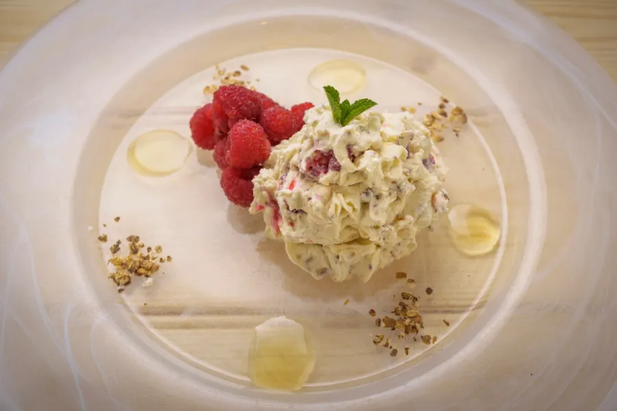 A portion of cranachan served on a plate with fresh raspberries and mint leaves with drops of honey and crumbled toasted oats as decoration.
